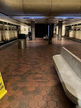 A picture of a train platform. To the left there is the bottom of a yellow wet floor cone and to the right there is a stone bench for sitting. In the background there are digital displays and pillars, as well as train tracks on the extreme left and right sides in the background. There is a ceiling at the top of the picture.